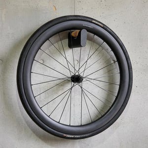 Könnte beinhalten: Schwarzes Fahrradrad mit dünnen Speichen, an der Wand befestigt. Der Reifen trägt die Aufschrift "GRAND PRIX 5000" und "ELITEWHEELS". Das Rad wird von einer schwarzen Halterung mit Karbonfasermuster gehalten.
