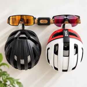 Könnte beinhalten: Ein wandmontiertes Holzregal mit einer schwarzen Halterung in der Mitte, die ein florales Logo zeigt. Zwei schwarze Haken halten einen schwarzen Fahrradhelm und einen weiß-rot-schwarzen Fahrradhelm. Zwei Sportbrillen liegen auf der Holzstange.