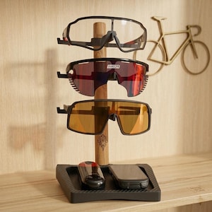 Può includere: Un espositore in legno per occhiali da sole con tre paia di occhiali sportivi con lenti di diversi colori. Un vassoio nero alla base contiene un piccolo dispositivo e uno smartphone. Una decorazione da parete a forma di bicicletta color oro è sullo sfondo.