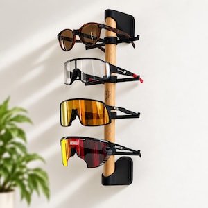 Könnte beinhalten: Ein vertikaler Wand-Brillenhalter mit einer zentralen Stange aus hellem Holz und schwarzen Kunststoffhalterungen. Das Regal bietet Platz für vier Brillen, darunter eine mit braunen Gläsern, eine mit klaren Gläsern und zwei mit orangefarbenen.