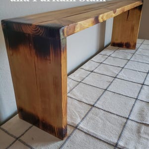 May include: A wooden bench with a partial wood burn and Puritan stain finish. The bench has a rectangular top and two supporting legs. The wood has a natural grain and a dark brown, burnt appearance on the edges. The text "Partial Wood Burn and Puritan Stain" is visible.