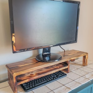 May include: A wooden monitor stand with a keyboard shelf. The stand is made of stained wood with visible grain. A black computer monitor sits on top, and a black keyboard rests on the shelf below. The stand is approximately 30 inches wide.
