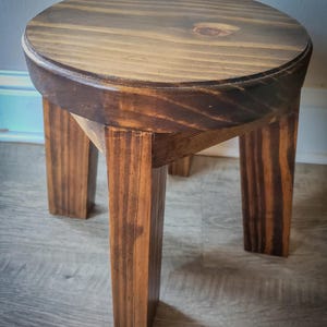 Könnte beinhalten: Ein kleiner, runder Holzschemel mit dunkelbraunem Anstrich. Der Hocker hat eine kreisförmige Sitzfläche und vier quadratische Beine. Die Holzmaserung ist auf Sitzfläche und Beinen sichtbar. Der Hocker ist etwa 30 cm hoch.