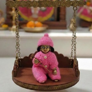 May include: A miniature brass swing holding a doll in pink knitted attire and a matching hat. The doll holds a small stick. The swing hangs by chains, set against religious icons and a wooden shrine, creating a devotional scene.