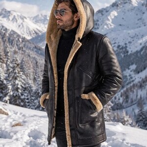 Könnte beinhalten: Ein schwarzer Ledercoat mit beigem Shearling-Futter und Kapuze. Der Mantel hat ein klassisches Design mit Knopfverschluss und Taschen. Die Person auf dem Bild trägt eine Sonnenbrille und steht in einer schneebedeckten Berglandschaft.