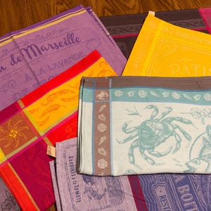 May include: A collection of colorful, folded kitchen towels with French text. The towels feature various designs, including a crab, fish, and text like "Savon de Marseille". Colors include purple, yellow, red, and teal. The towels are made of a woven fabric.
