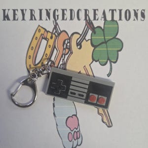 May include: A grey keychain with a miniature replica of a retro video game controller. The controller has black buttons and a directional pad. The keychain is attached to a silver clasp. The text "KEYRINGEDCREATIONS" is at the top.