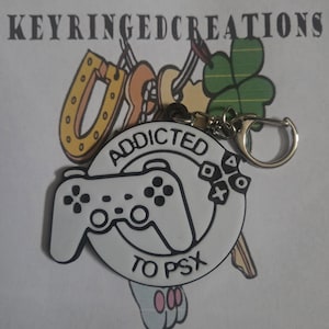 May include: A white and black keychain with the text "ADDICTED TO PSX" and a controller graphic. The keychain has a silver clasp and is attached to other charms, including a horseshoe and a four-leaf clover.