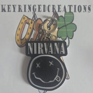 May include: A black and white Nirvana smiley face keyring. The keyring features the word "NIRVANA" in white block letters above the smiley face. It is attached to a silver keyring.