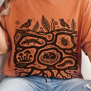 May include: A burnt orange short-sleeve t-shirt featuring a black graphic print of a cross-section of earth. The design includes a mouse, a beetle, a lizard, mushrooms, a centipede, ferns, and two birds. The word Yam is printed in the top right corner.