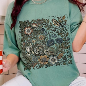 May include: Light green t-shirt featuring a detailed floral and fauna design. The design includes a cat, bird, and various flowers and leaves in shades of brown, blue, and green. The shirt has short sleeves and a relaxed fit. The words "LIGHT GREEN" are in a circle in the upper left corner.