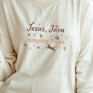 Può includere: Felpa a maniche lunghe color avorio con la scritta "Jesus, Java Jumping Paws" in marrone. Il design include impronte di zampe, tazze di caffè, un cane che salta e una croce. La felpa ha un girocollo e una vestibilità ampia.