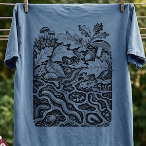 May include: A blue jean coloured t-shirt featuring a dark blue graphic print of a forest floor ecosystem. The illustration depicts various insects, worms, mushrooms, and oak leaves in a cross-section view of soil. The text Blue Jean is visible.