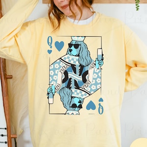 May include: A light yellow sweatshirt featuring a playing card design with a dog wearing sunglasses and a crown, holding a drink. The card is outlined in black with blue and white details. The word "BUTTER" is in a circle at the bottom.
