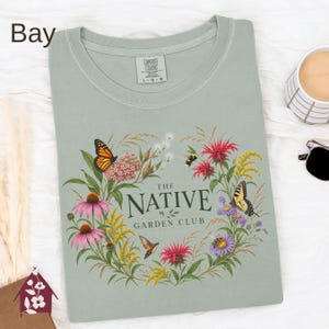 May include: A sage green t-shirt featuring a graphic of a floral wreath with butterflies, a bee, and a hummingbird. The text in the center reads THE NATIVE GARDEN CLUB. The shirt has a crew neckline and a Comfort Colors brand label at the inner neck.
