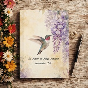 May include: A journal with a watercolor illustration of a hummingbird and wisteria. The journal cover has the text "He makes all things beautiful, Ecclesiastes 3:11". A pen rests on the wooden surface next to the journal.