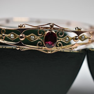 May include: A gold-coloured bracelet with a central oval, deep red gemstone. The bracelet has detailed filigree and small, round pearls. The design incorporates diamond and swirl patterns. The bracelet is shown on a dark green and black surface.
