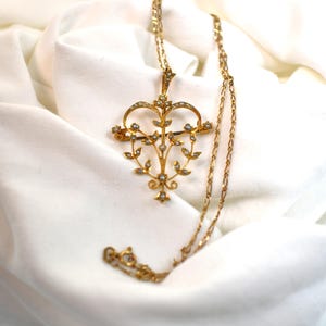 May include: A gold pendant necklace with a heart-shaped design and floral and leaf detailing. The pendant is embellished with small, round, pearl-like elements. The necklace chain is also gold and delicate in appearance.