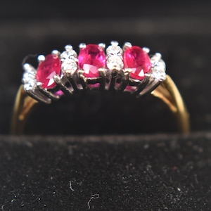 May include: A gold ring featuring five oval-cut, vibrant red gemstones, likely rubies, set in a silver-colored band. Small, clear accent stones are interspersed between the rubies. The ring is displayed in a black jewelry box.