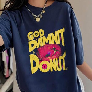 Może przedstawiać: Granatowa koszulka z żółtą grafiką z napisem "GOD DAMNIT DONUT". Grafika przedstawia scenę z kreskówki z postaciami i kształtem pączka. Koszulka ma krótkie rękawy i okrągły dekolt.