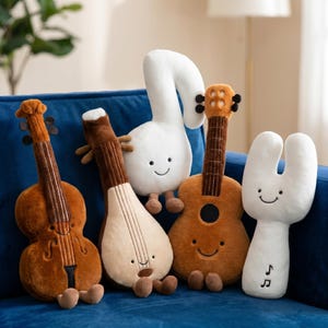 Puede incluir: Una colección de instrumentos musicales de peluche, incluyendo un violín, un laúd, una guitarra y dos figuras con forma de notas musicales. Los peluches son en tonos marrones y blancos, con caras sonrientes. Están colocados sobre una superficie azul.