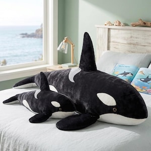 Peut inclure: Deux coussins en forme d'orques, un grand et un petit, sont présentés sur un lit. Les baleines sont noires et blanches, avec des détails précis. Un livre sur la vie marine est ouvert sur le lit.