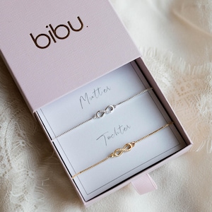 Peut inclure: Une boîte cadeau rose avec le nom de la marque "bibu" imprimé sur le couvercle. À l'intérieur, deux bracelets délicats avec le symbole de l'infini sont présentés, un argenté et un doré, avec les mots "Mutter" et "Tochter" écrits sur la carte.