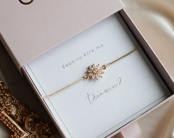 May include: A gold-colored bracelet with a flower-shaped pendant, presented in a pink box. The pendant is adorned with clear crystals. The box contains the text "Veux-tu être ma témoin?" in French.