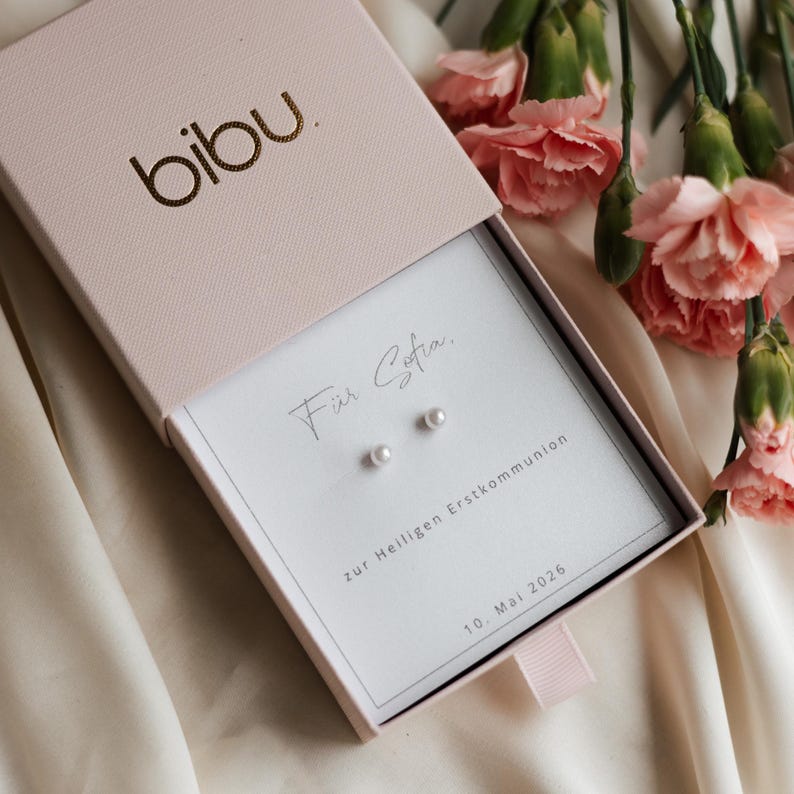 May include: A light pink jewelry box with the brand name "bibu" in gold lettering. Inside, a white card displays two small pearl earrings and the text "F&uuml;r Sofia" and "zur Heiligen Erstkommunion 10. Mai 2026". Pink carnations are in the background.