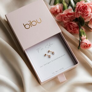 May include: A light pink jewelry box with the gold-colored brand name "bibu" on the lid. Inside, a pair of gold earrings with small, white pearls are displayed on a white card. The card has text in script and block letters. Pink carnations are in the background.