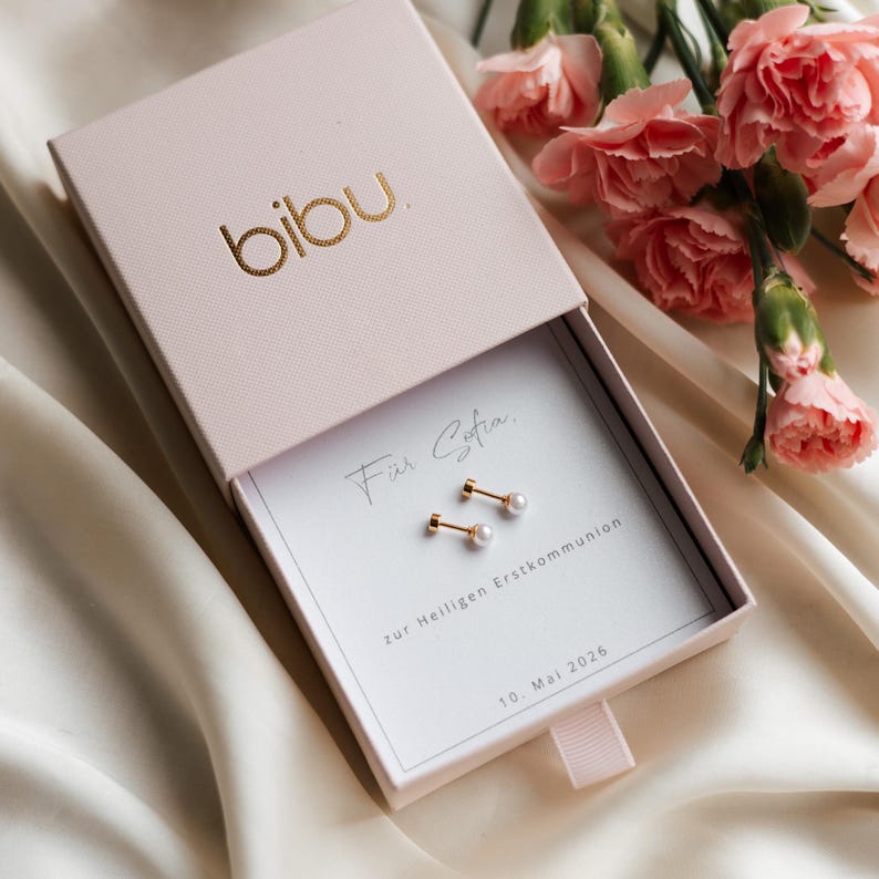 May include: A light pink jewelry box with the gold-colored brand name "bibu" on the lid. Inside, a pair of gold earrings with small, white pearls are displayed on a white card. The card has text in script and block letters. Pink carnations are in the background.