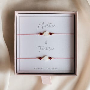 Può includere: Un set di due braccialetti coordinati con cordino rosa, perline bianche a forma di cuore e dettagli rossi, presentati in una scatola regalo rosa. La scatola presenta le parole "Mutter & Tochter" e il marchio "rubin · perlmutt".
