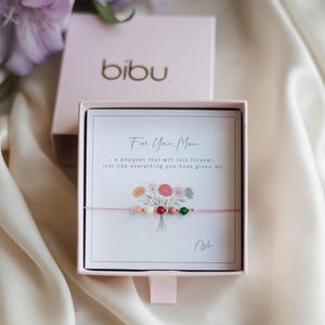 Könnte beinhalten: Eine rosa Geschenkbox mit dem Markennamen "bibu" auf dem Deckel. Im Inneren befindet sich eine Karte mit der Aufschrift "For You, Mom" mit einer floralen Illustration und einem Perlenarmband. Das Armband hat rosa, rote und grüne Perlen.