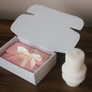 May include: A white gift box with an open lid, revealing a pink envelope tied with a cream ribbon. The ribbon has a small tag with the word "bibu". A white, layered candle sits to the right of the box on a wooden surface.