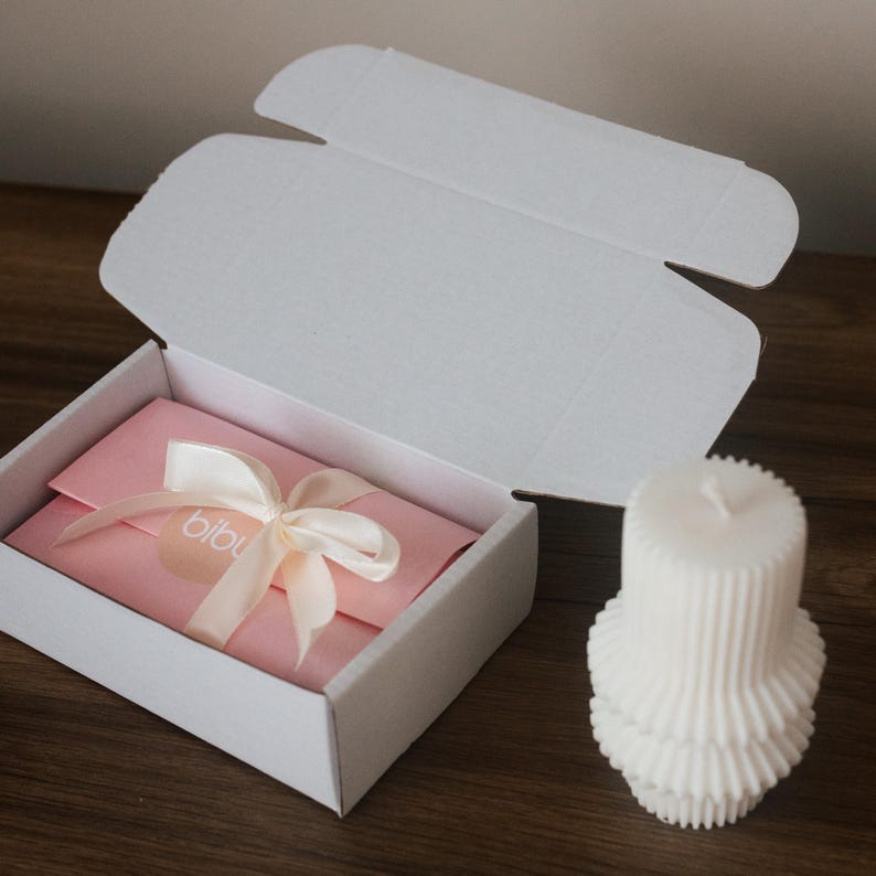 May include: A white gift box with an open lid, revealing a pink envelope tied with a cream ribbon. The ribbon has a small tag with the word "bibu". A white, layered candle sits to the right of the box on a wooden surface.