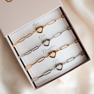 Op de afbeelding: Een set van vier paperclip kettingarmbanden in een roze geschenkdoos. Twee zijn goudkleurig en twee zilverkleurig. Elke armband heeft een hartvormige knoop. De doos is gelabeld met de woorden "Mom", "Dad", "Daughter" en "Son".