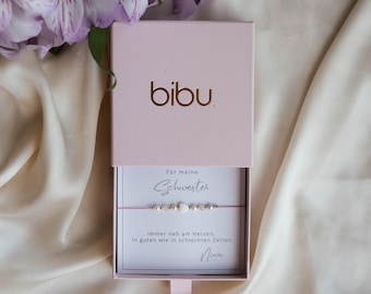 May include: A pink gift box with the brand name "bibu" in gold lettering. Inside, a bracelet with white beads and gold accents rests on a card with German text. The box is open, revealing the bracelet and card. Lilac flowers are in the background.