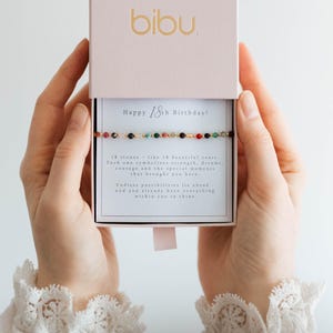Op de afbeelding: Een goudkleurige armband met kleurrijke kralen wordt gepresenteerd in een roze doos. Op het deksel van de doos staat de merknaam "bibu" in goud. De kaart binnenin luidt "Happy 18th Birthday!" en beschrijft de symboliek van de armband.