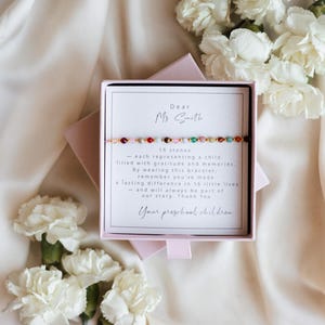 Könnte beinhalten: Eine rosa Geschenkbox mit einem Armband und einer Karte. Die Karte ist an "Ms Smith" adressiert und beschreibt ein Armband mit 16 Steinen, die jeweils ein Kind repräsentieren. Das Armband hat bunte Perlen. Weiße Blumen sind im Hintergrund.