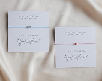 May include: Two friendship bracelets on cards. One bracelet is light blue with a light blue bead and gold accents. The other is pink with a red bead and gold accents. The cards read "I couldn't imagine anyone better... Will you be my Godmother?"