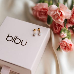 May include: A pair of gold stud earrings with white pearls, displayed on a light pink box with the word "bibu" printed on it. Pink carnation flowers are in the background, with a white satin cloth.
