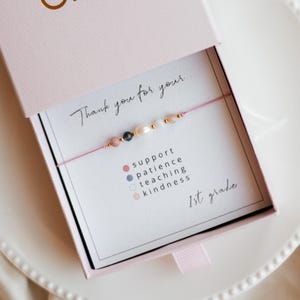 Könnte beinhalten: Eine rosa Geschenkbox mit einem Armband und einer Karte mit der Aufschrift "Vielen Dank für Ihre...". Das Armband hat Perlen und Perlen. Die Karte listet "Unterstützung, Geduld, Lehre, Freundlichkeit" und "1. Klasse" auf.