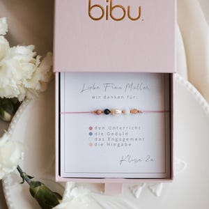 May include: A pink gift box with the gold text "bibu" on the lid. Inside, a pink string bracelet with beads is displayed on a card with German text. The card reads "Liebe Frau Müller, wir danken für:" and lists qualities.