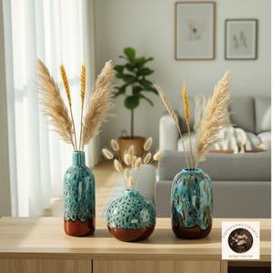 Puede incluir: Tres jarrones de cerámica con arreglos florales secos. Los jarrones tienen un esmalte turquesa y marrón. El jarrón más alto tiene forma de botella, mientras que los otros dos son más redondos. Los jarrones se exhiben sobre una superficie de madera.