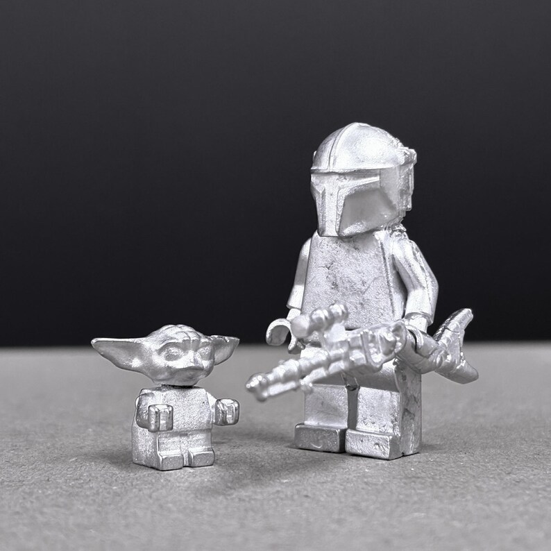 May include: Two silver-colored figurines: a Mandalorian figure with a blaster and a small Grogu figure. The Mandalorian figure is wearing a helmet and armor. The Grogu figure has large ears and is in a standing position. The figures are on a gray surface.