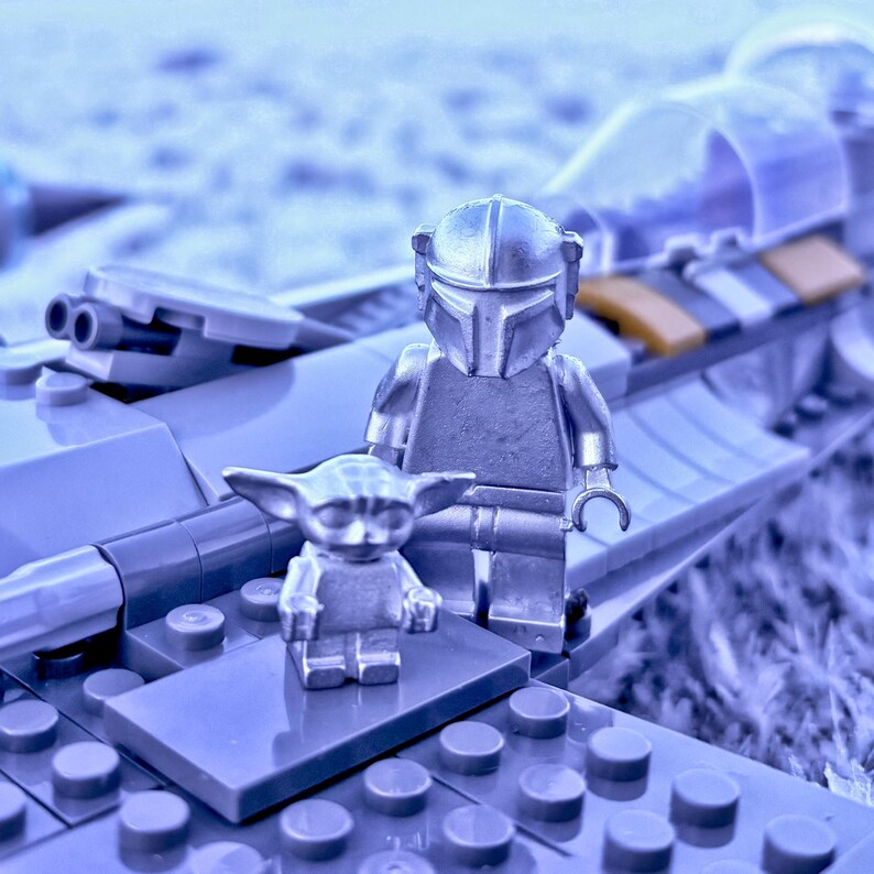 May include: A silver-colored Lego set featuring two figures: a Mandalorian and a Grogu. The figures stand on a gray Lego base, with a partially visible spaceship in the background. The scene is set against a soft, blurred backdrop.