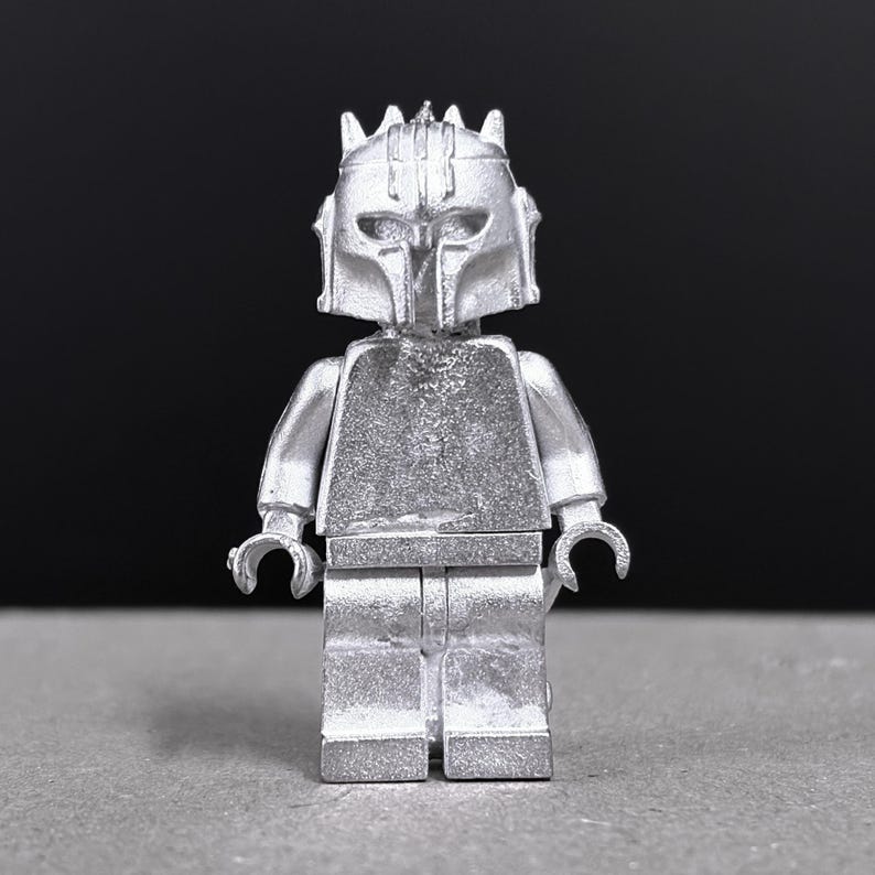 May include: A silver-colored Lego minifigure with a helmet and armor. The figure has a detailed design, including a helmet with a visor and a chest plate. The arms and legs are articulated, and the figure is standing on a gray surface.