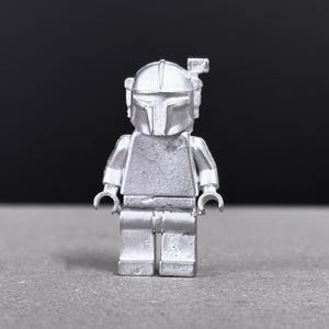May include: A silver-colored Lego figure of a Mandalorian warrior, featuring a helmet with a T-shaped visor and a small antenna. The figure is standing on a gray surface with a black background.