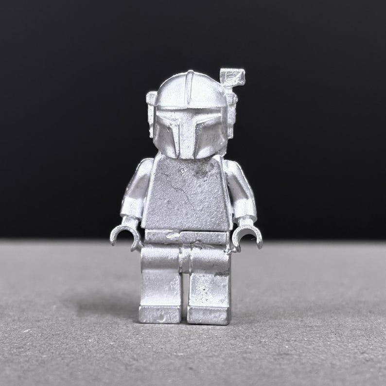 May include: A silver-colored Lego figure of a Mandalorian warrior, featuring a helmet with a T-shaped visor and a small antenna. The figure is standing on a gray surface with a black background.