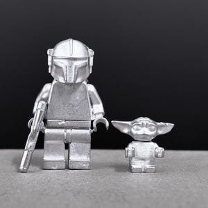 May include: Two silver-colored Lego figures: a Mandalorian with a helmet and blaster, and a small Grogu figure. The figures are on a gray surface with a black background. The Mandalorian figure is approximately 4 inches tall.